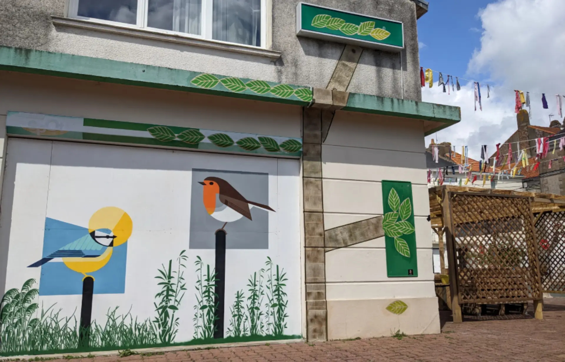 Décoration murale extérieure d'un bâtiment avec des feuilles peintes en vert et deux oiseaux, un rouge-gorge familier et une mésange bleue, sur un fond blanc.